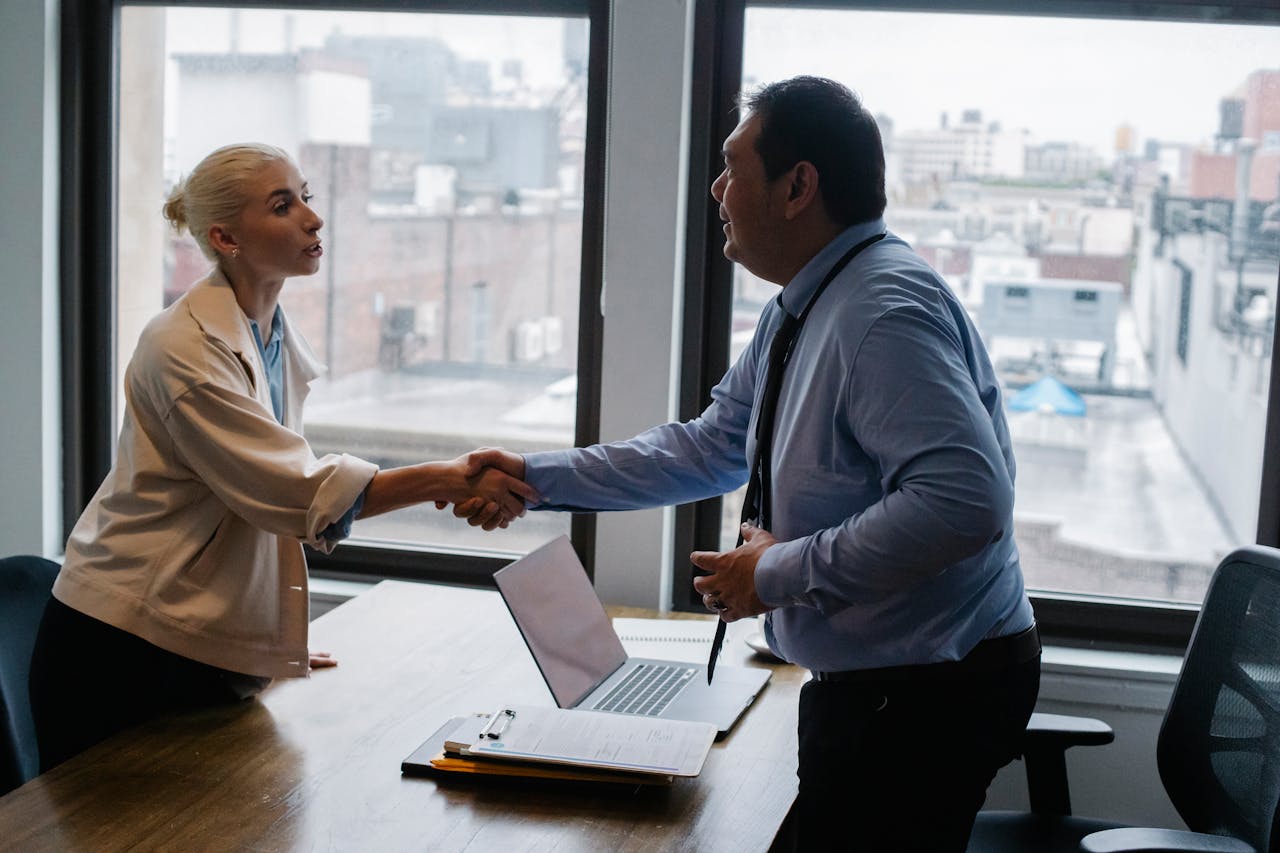 Young female candidate shaking hands with employer before job interview in light office with urban landscape through windows