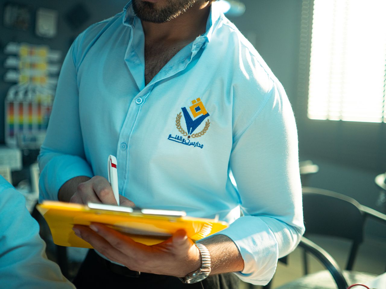 who-we-are Man in professional attire holding a yellow notepad and pen in an office setting, depicted clearly with branding.