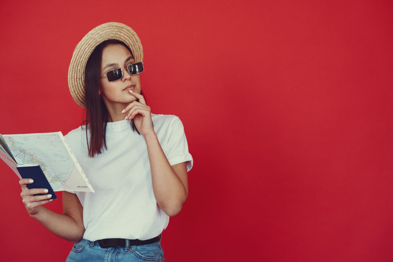 who-we-are Thoughtful young woman in retro hat and trendy sunglasses holding map and touching chin while looking pensively away on red background