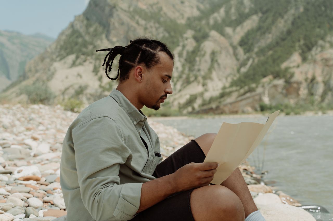 A man sits by a river, reading a map amidst scenic mountain views, embodying adventure and exploration.
